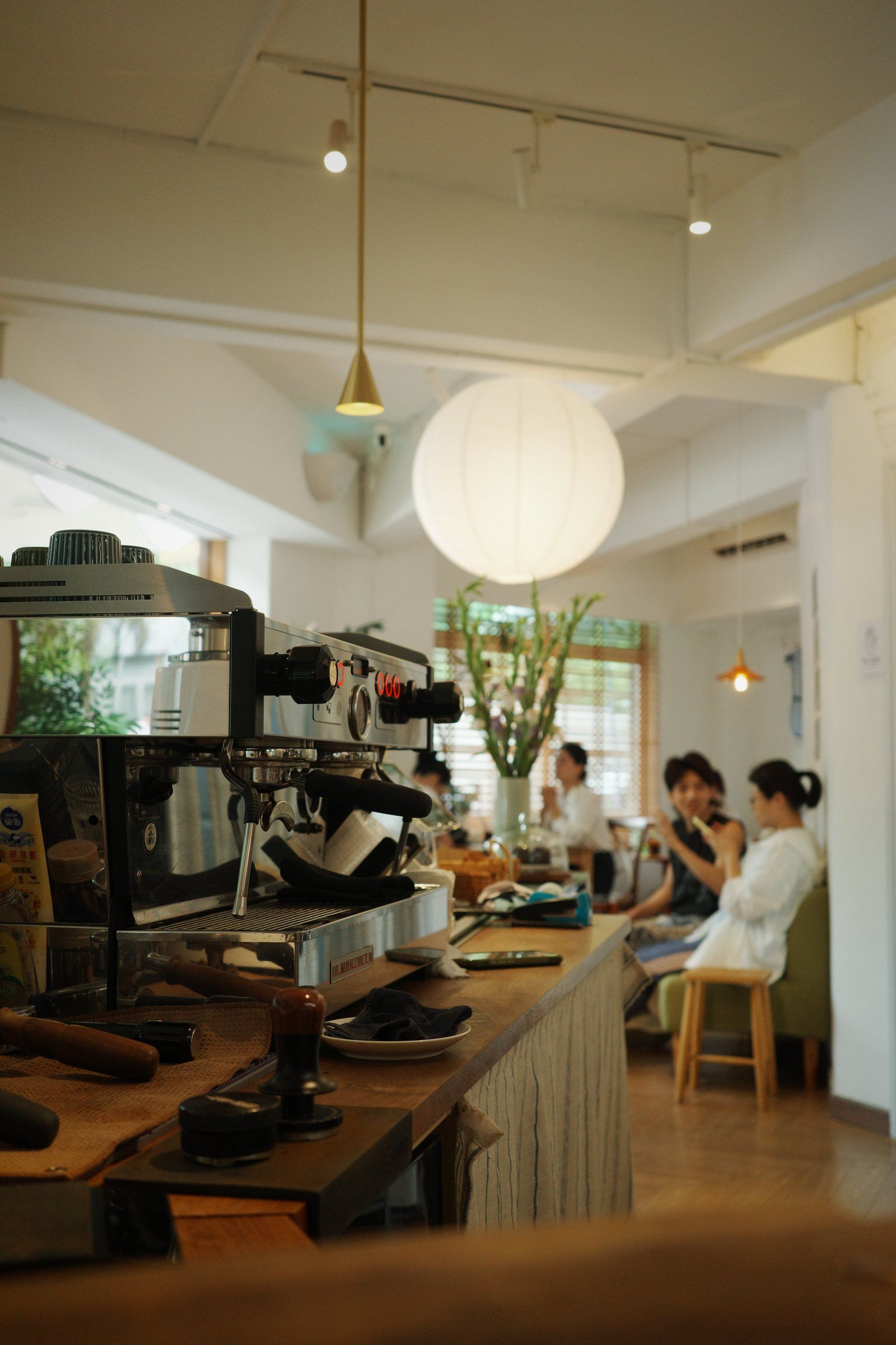 店内咖啡机与木质吧台在前景，后方有顾客坐在灯光下